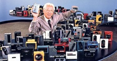 Akio Morita, cofondateur de Sony, souriant au milieu d'une vaste collection de modèles de Walkman vintage, tenant un prototype et un baladeur cassette.