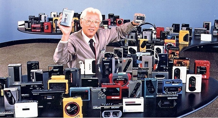 Akio Morita, cofondateur de Sony, souriant au milieu d'une vaste collection de modèles de Walkman vintage, tenant un prototype et un baladeur cassette.