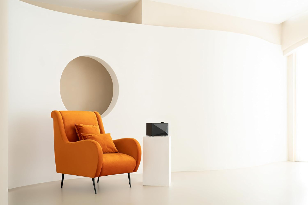 Enceinte Bluetooth Astell & Kern Acro BE100 noire posée sur un piédestal blanc dans un salon minimaliste à côté d'un fauteuil orange design.