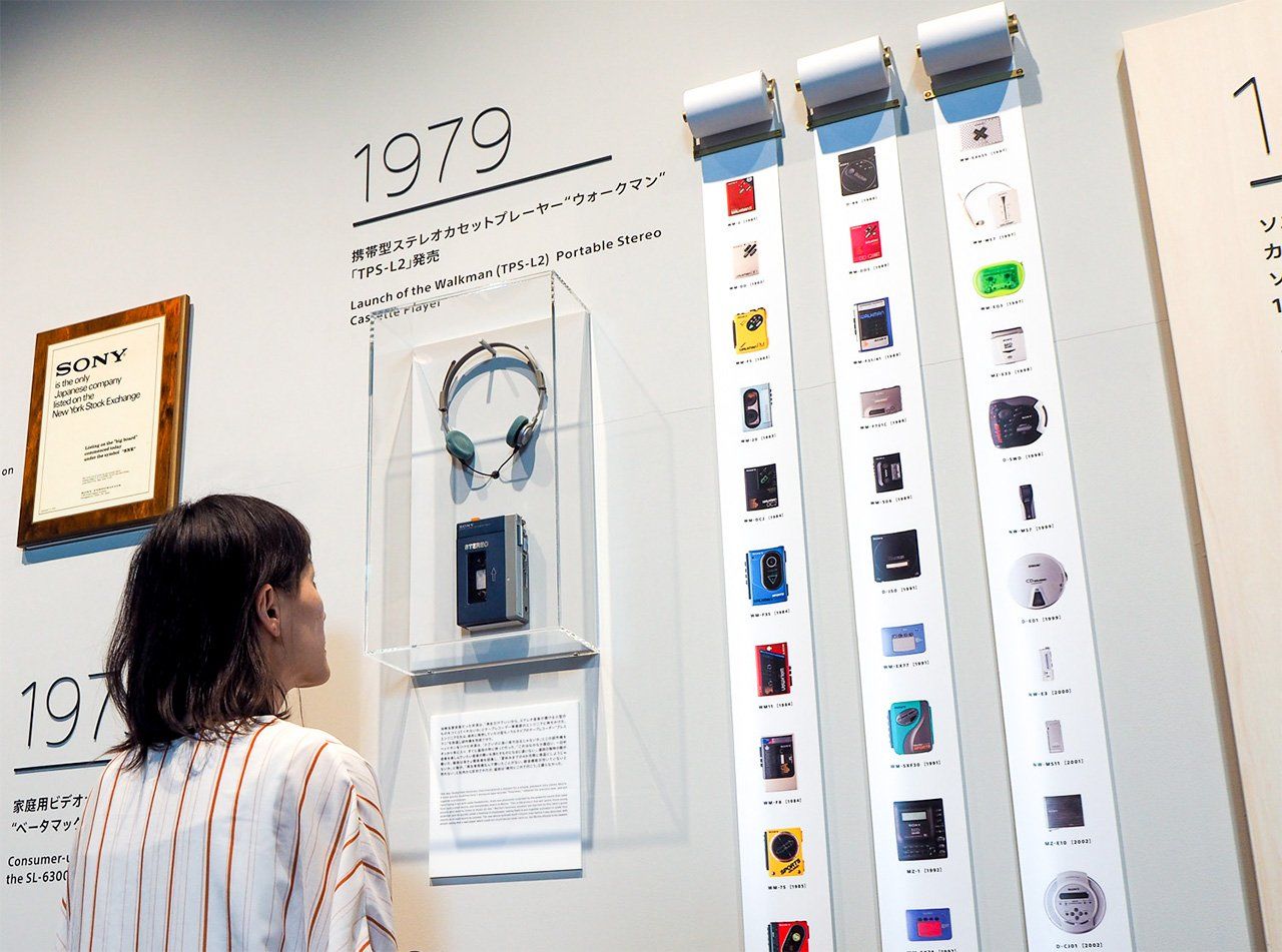 Une visiteuse devant un mur d'exposition Sony présentant le premier Walkman TPS-L2 de 1979 sous vitrine et des colonnes listant les nombreux modèles dérivés des années 80 et 90.