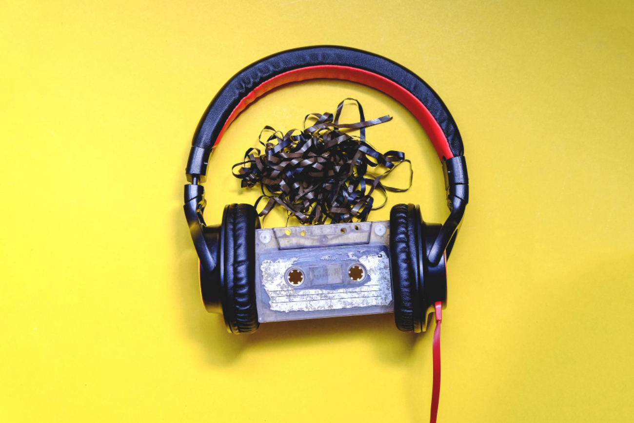 Gros plan d'un casque filaire noir et rouge entourant une cassette audio usée avec sa bande magnétique emmêlée sur fond jaune.