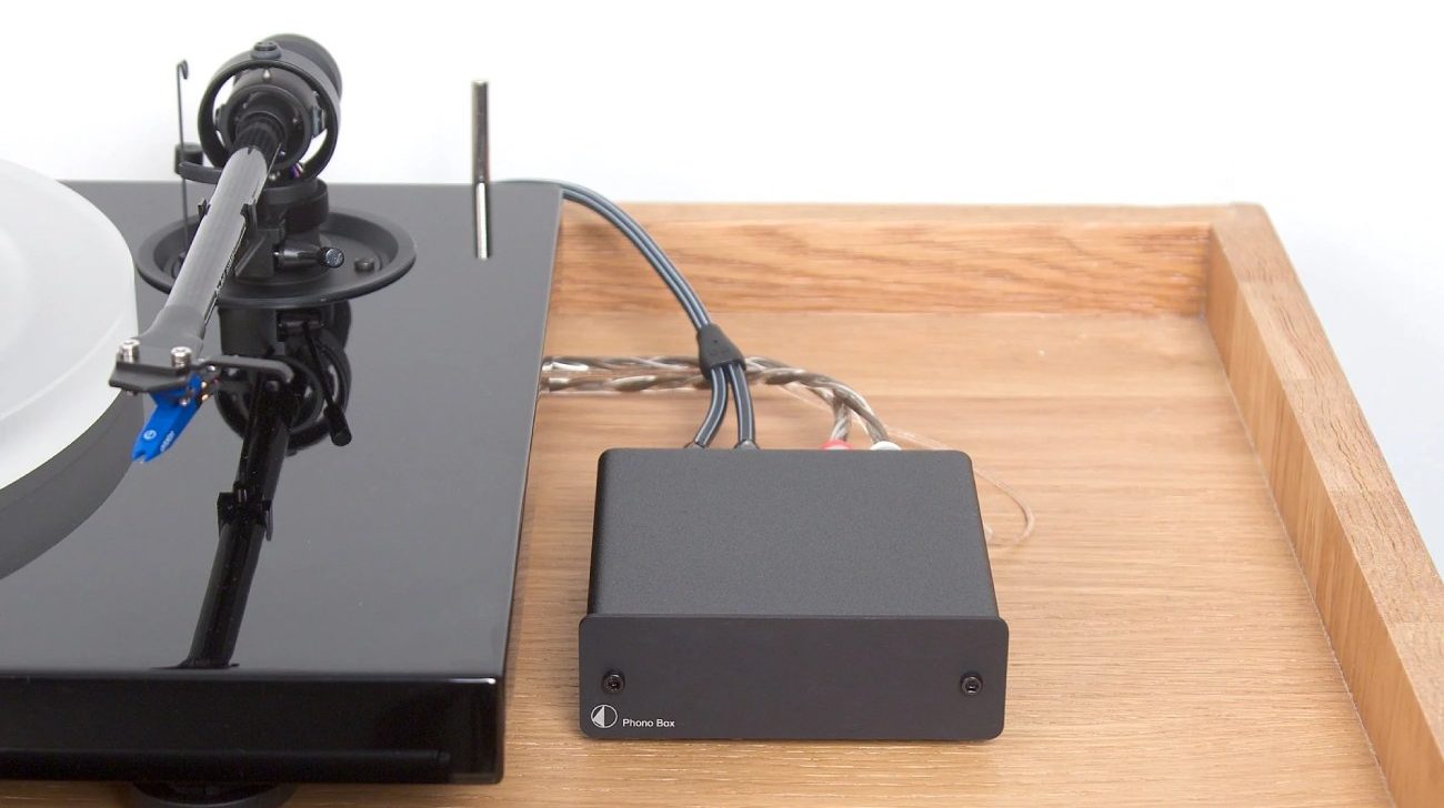 Mise en situation d'un préamplificateur Pro-Ject Phono Box noir relié à une platine vinyle Pro-Ject avec cellule Ortofon Blue sur un meuble en bois.