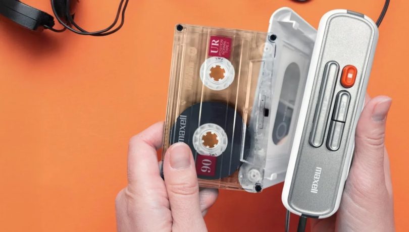 Vue de dessus du lecteur cassette portable Maxell MXCP-P100 blanc et gris, ouvert, avec une cassette Maxell UR 90 insérée sur un fond orange vif.
