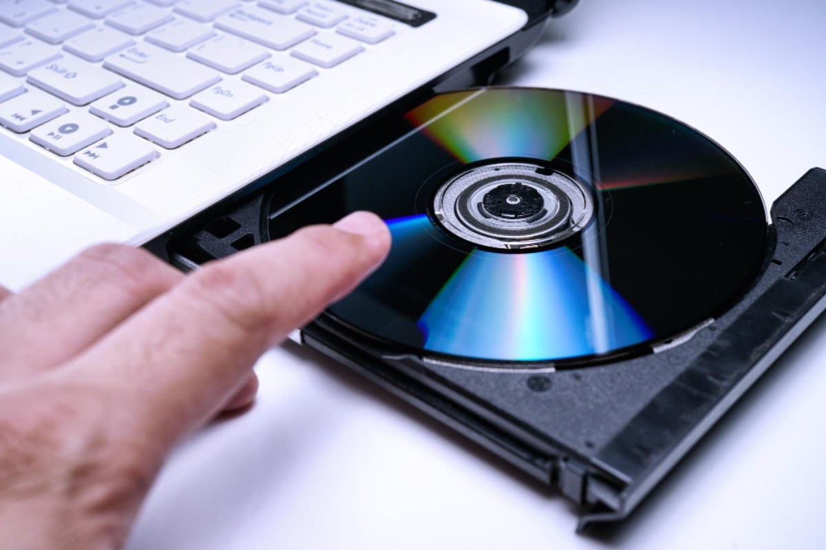 Un gros plan sur un tiroir de lecteur CD de laptop ouvert avec une main s'apprêtant à insérer ou retirer un disque, illustrant le processus de gravure et le concept d'overburning vers le bord du disque.