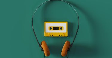 Vue de dessus d'un casque audio filaire vintage à arceau fin et mousses orange entourant une cassette audio jaune sur fond vert émeraude.