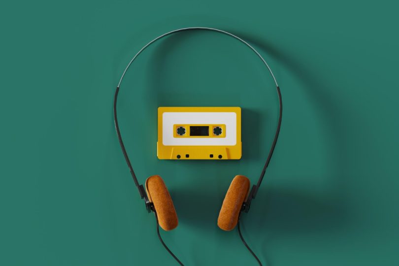 Vue de dessus d'un casque audio filaire vintage à arceau fin et mousses orange entourant une cassette audio jaune sur fond vert émeraude.