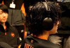 Photo de dos d'un joueur d'esport portant le casque Sony InZone H9 II noir. Sa chevelure texturée et l'oreillette noire mate sont bien visibles, avec en arrière-plan flou un coéquipier portant le maillot de l'équipe Fnatic avec des accents orange.