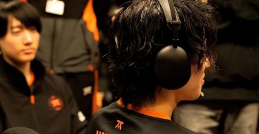 Photo de dos d'un joueur d'esport portant le casque Sony InZone H9 II noir. Sa chevelure texturée et l'oreillette noire mate sont bien visibles, avec en arrière-plan flou un coéquipier portant le maillot de l'équipe Fnatic avec des accents orange.