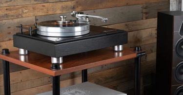 Platine vinyle VPI Forever Model One avec châssis en chêne et plateau en aluminium, équipée du bras en S sur un support dédié, posée sur un meuble hi-fi à côté d'une enceinte colonne.