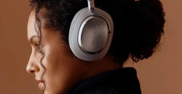 Jeune femme portant le casque sans fil Bowers & Wilkins Px8 S2 en version beige clair avec coques métalliques brossées, fond uniformément coloré terre cuite