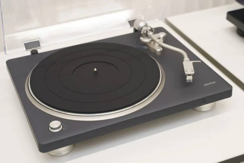 Platine vinyle Denon DP-500BT avec capot plastique transparent ouvert, plateau en caoutchouc noir, bras tangentiel argenté et pieds de découplage chromés
