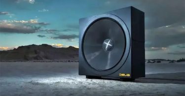Subwoofer infrasonique Deus Audio Invicta 60 photographié dans un paysage désertique au coucher du soleil, châssis cubique imposant noir mat avec membrane circulaire de 60 pouces de diamètre dotée d'un dôme central orné d'un X chromé, logo DEUS jaune doré gravé en bas à droite, monolithe technologique posé seul sur un sol de sable