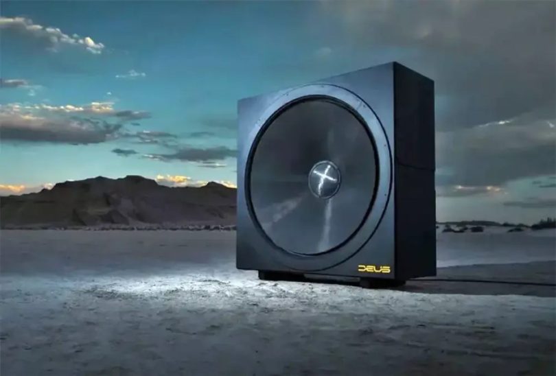 Subwoofer infrasonique Deus Audio Invicta 60 photographié dans un paysage désertique au coucher du soleil, châssis cubique imposant noir mat avec membrane circulaire de 60 pouces de diamètre dotée d'un dôme central orné d'un X chromé, logo DEUS jaune doré gravé en bas à droite, monolithe technologique posé seul sur un sol de sable