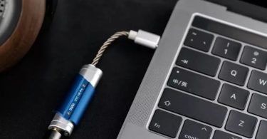 Dongle DAC USB portable DUNU DTC100 Pro en finition bleu et argent connecté à un MacBook Pro via USB-C, avec casque audio en arrière-plan sur tapis noir
