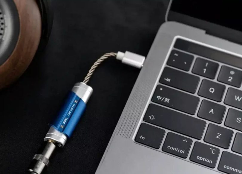 Dongle DAC USB portable DUNU DTC100 Pro en finition bleu et argent connecté à un MacBook Pro via USB-C, avec casque audio en arrière-plan sur tapis noir