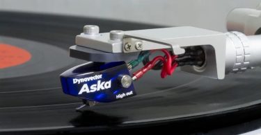 Cellule phono moving coil Dynavector Aska version High Out en bleu translucide, montée sur un bras de lecture au-dessus d'un vinyle, avec cantilever aluminium et stylus elliptique