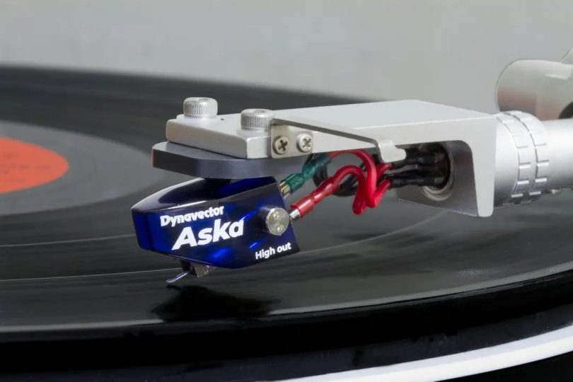Cellule phono moving coil Dynavector Aska version High Out en bleu translucide, montée sur un bras de lecture au-dessus d'un vinyle, avec cantilever aluminium et stylus elliptique
