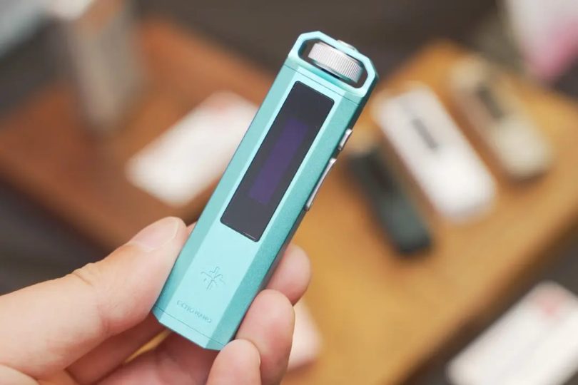Lecteur audio numérique FiiO Snowsky ECHO NANO en finition turquoise tenu dans une main, avec écran OLED noir et molette de navigation argentée en haut, sur fond de bois flou