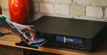 Lecteur CD NAD C 589 avec tiroir ouvert et disque inséré, afficheur numérique rétroéclairé, installé sur un meuble bois devant un mur de briques apparentes