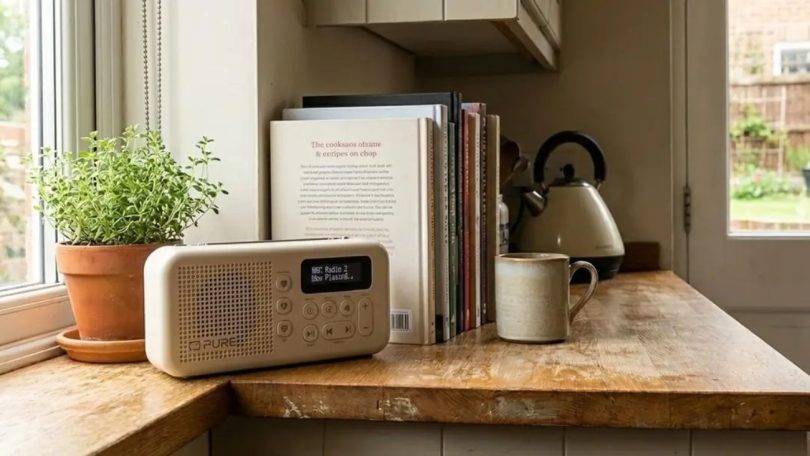 Radio portable Pure Elan Move blanche posée sur un plan de travail en bois près d'une fenêtre de cuisine, avec plante aromatique, livres de cuisine et tasse en céramique