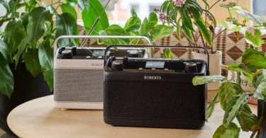 Duo de radios portables Roberts Stream 219 et Blutune 7 posées sur une table en bois entourée de plantes vertes, versions blanche et noire côte à côte avec antenne télescopique déployée
