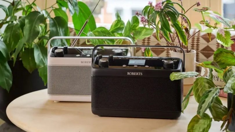 Duo de radios portables Roberts Stream 219 et Blutune 7 posées sur une table en bois entourée de plantes vertes, versions blanche et noire côte à côte avec antenne télescopique déployée