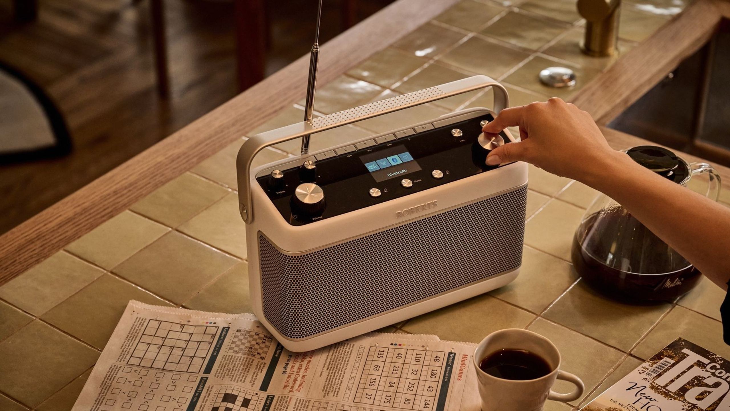 Roberts Stream 219 blanc posé sur une table de cuisine en carrelage, antenne télescopique déployée, écran affichant le mode Bluetooth, à côté d'une tasse de café et d'un journal ouvert aux mots croisés