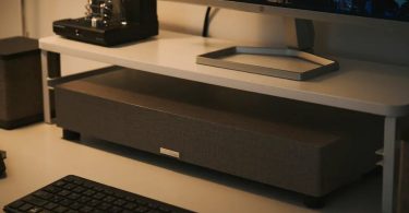 Subwoofer SOUND WARRIOR SW-W1 ultra-fin de 7 cm posé sous un écran PC HP avec amplificateur à tubes visible en arrière-plan, dans un setup bureau audiophile en ambiance tamisée