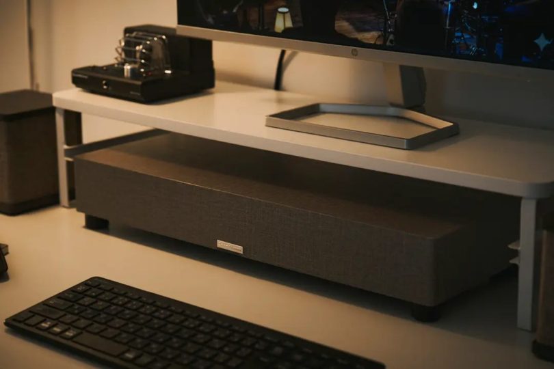 Subwoofer SOUND WARRIOR SW-W1 ultra-fin de 7 cm posé sous un écran PC HP avec amplificateur à tubes visible en arrière-plan, dans un setup bureau audiophile en ambiance tamisée