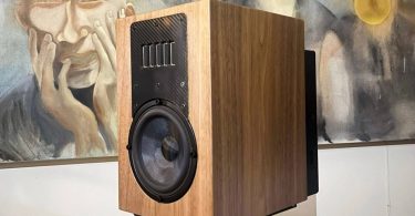 Prototype d'enceinte Acoustic3D DaS en finition noyer naturel posée sur stand noir, baffle avant en fibre de carbone twill avec array vertical de drivers tweeter en partie haute et woofer médium-grave à cône argenté en partie basse, devant deux peintures de moines bouddhistes
