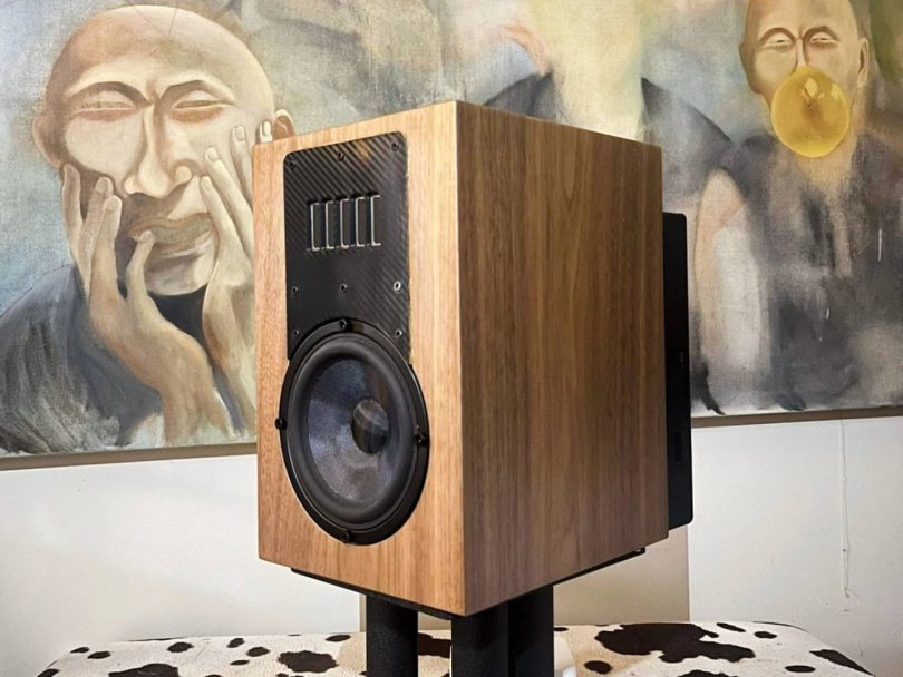 Prototype d'enceinte Acoustic3D DaS en finition noyer naturel posée sur stand noir, baffle avant en fibre de carbone twill avec array vertical de drivers tweeter en partie haute et woofer médium-grave à cône argenté en partie basse, devant deux peintures de moines bouddhistes
