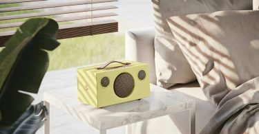 Enceinte connectée multiroom Audio Pro C5 MkII W Classic en édition limitée finition jaune vif avec poignée en cuir véritable couleur cognac sur le dessus, panneau de commande et grilles tweeter et woofer en métal couleur laiton brass, posée sur table en marbre blanc dans un salon lumineux moderne avec canapé beige clair et plante verte tropicale en arrière-plan