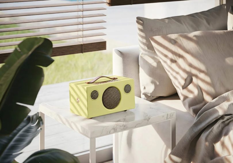Enceinte connectée multiroom Audio Pro C5 MkII W Classic en édition limitée finition jaune vif avec poignée en cuir véritable couleur cognac sur le dessus, panneau de commande et grilles tweeter et woofer en métal couleur laiton brass, posée sur table en marbre blanc dans un salon lumineux moderne avec canapé beige clair et plante verte tropicale en arrière-plan