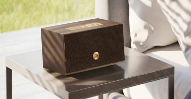 Enceinte connectée multiroom Audio Pro C5 MkII W en finition placage noyer véritable walnut veneer avec grille frontale en tissu marron foncé harmonisée au bois et bouton métallique laiton brass au centre, posée sur table d'appoint en marbre gris foncé dans un salon contemporain lumineux avec fauteuil beige clair et stores en bois lamellé filtrant la lumière naturelle, vue trois-quarts mise en scène lifestyle scandinave moderne