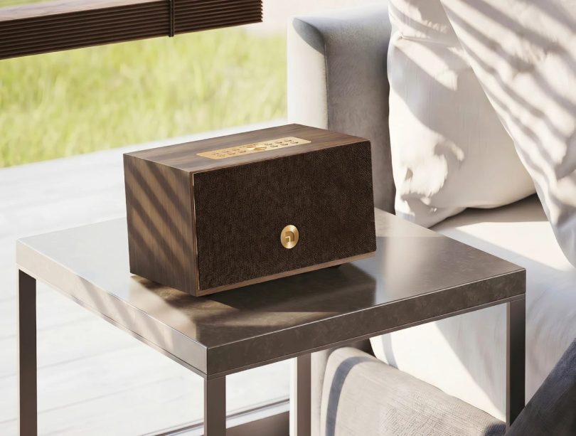 Enceinte connectée multiroom Audio Pro C5 MkII W en finition placage noyer véritable walnut veneer avec grille frontale en tissu marron foncé harmonisée au bois et bouton métallique laiton brass au centre, posée sur table d'appoint en marbre gris foncé dans un salon contemporain lumineux avec fauteuil beige clair et stores en bois lamellé filtrant la lumière naturelle, vue trois-quarts mise en scène lifestyle scandinave moderne
