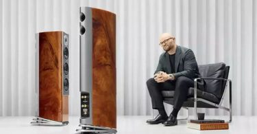 Paire d'enceintes colonne Audiovector R5 Arreté en finition placage acajou africain laqué piano avec veinages chauds, à côté de Mads Klifoth CEO d'Audiovector assis dans un fauteuil en cuir noir, dans un studio d'écoute aux rideaux blancs plissés