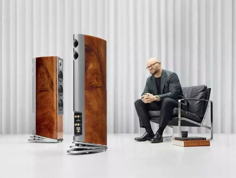 Paire d'enceintes colonne Audiovector R5 Arreté en finition placage acajou africain laqué piano avec veinages chauds, à côté de Mads Klifoth CEO d'Audiovector assis dans un fauteuil en cuir noir, dans un studio d'écoute aux rideaux blancs plissés