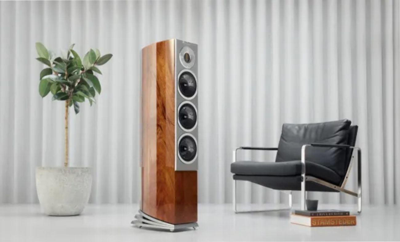 Enceinte colonne Audiovector R5 Arreté en finition African Mahogany Piano vue de face, montrant la configuration 3.5 voies : tweeter AMT chromé avec acoustic lens, driver médium dédié et deux haut-parleurs de grave 6,5 pouces en carbon-sandwich, plinthe Arreté en aluminium poli, dans un studio d'écoute avec fauteuil cuir noir et plante verte
