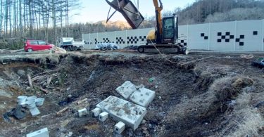 Vue du chantier du studio SKYSQUARE d'AZSTOKE à Yamanashi : pelleteuse jaune en action au-dessus de la fosse Foley en cours d'excavation, blocs de béton préparés au sol pour les enregistrements high-energy, mur d'isolation acoustique à damiers en arrière-plan