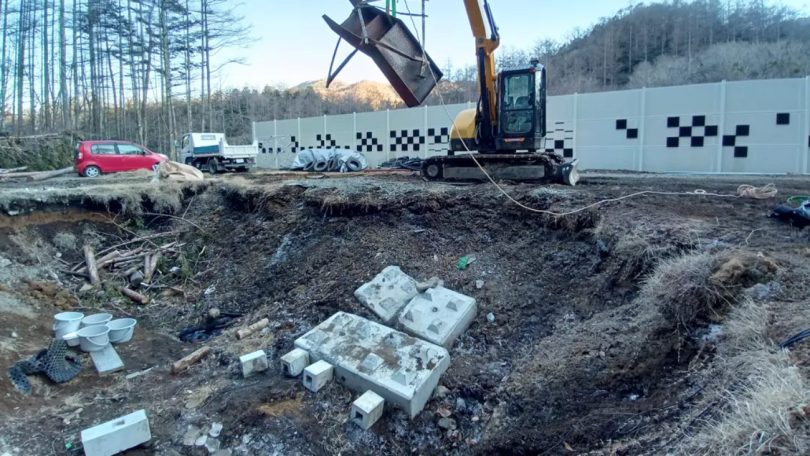 Vue du chantier du studio SKYSQUARE d'AZSTOKE à Yamanashi : pelleteuse jaune en action au-dessus de la fosse Foley en cours d'excavation, blocs de béton préparés au sol pour les enregistrements high-energy, mur d'isolation acoustique à damiers en arrière-plan
