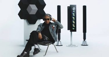 Hiroshi Fujiwara fondateur de Fragment Design assis avec le casque Beoplay H100 sur les oreilles, entouré de la collection Bang & Olufsen × Fragment Design : Beosound Shape en configuration fleur à gauche, Beosystem 9000c trois colonnes à lecteur CD vertical au centre et enceintes colonnes Beolab à droite, toutes en finition aluminium anodisé noir gloss