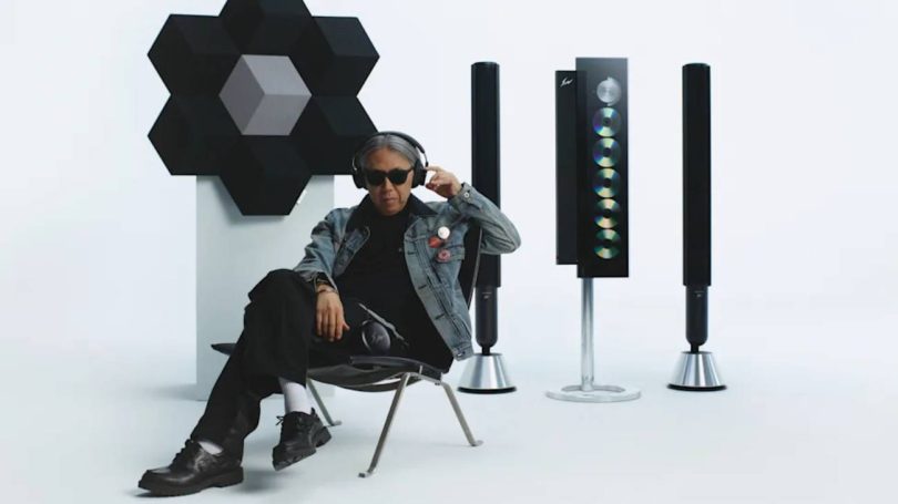 Hiroshi Fujiwara fondateur de Fragment Design assis avec le casque Beoplay H100 sur les oreilles, entouré de la collection Bang & Olufsen × Fragment Design : Beosound Shape en configuration fleur à gauche, Beosystem 9000c trois colonnes à lecteur CD vertical au centre et enceintes colonnes Beolab à droite, toutes en finition aluminium anodisé noir gloss