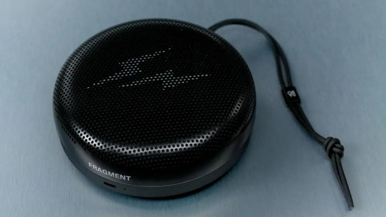 Enceinte portable Bang & Olufsen Beosound A1 3rd Gen en édition Fragment Design, finition aluminium anodisé noir gloss liquid-like, logo double éclair sous la grille perforée, dragonne en cuir noir tressé