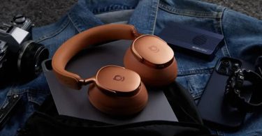 Casque circum-aural Baseus Inspire XH1 en coloris Orange Aurore aux coupelles cuivrées posé sur un blouson en jean, entouré d'un appareil photo argentique, d'une enceinte portable et d'une console de jeu