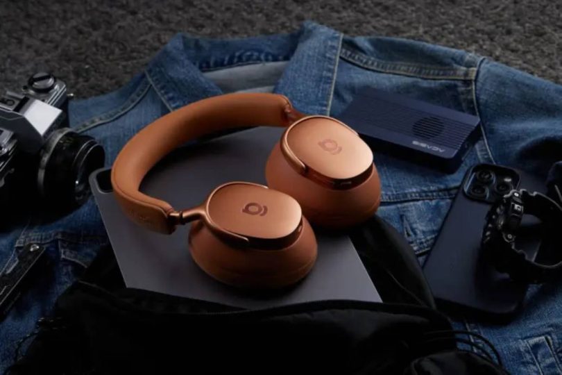 Casque circum-aural Baseus Inspire XH1 en coloris Orange Aurore aux coupelles cuivrées posé sur un blouson en jean, entouré d'un appareil photo argentique, d'une enceinte portable et d'une console de jeu