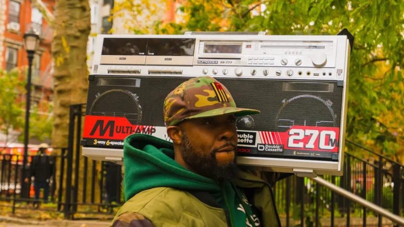 Homme portant un boombox Bumpboxx BB-777 sur l'épaule dans une rue de New York, design rétro inspiré du Sharp GF-777 de 1981 avec marquages Multi-Amp 3-way 6-speaker, Double Cassette et 270W