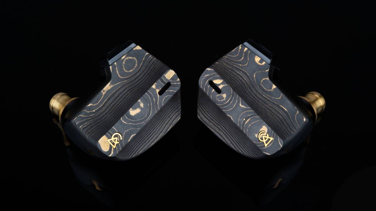 Paire d'écouteurs Campfire Audio Chimera vue de face sur fond noir, faceplates Damascus carbone-laiton aux motifs uniques avec couches de laiton dorées plongeant dans la fibre de carbone, nozzles en laiton usiné CNC et logo Campfire Audio en or