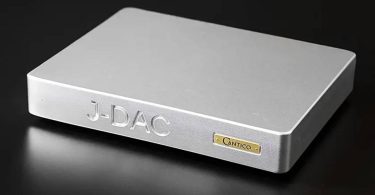Vue trois-quarts avant du convertisseur numérique-analogique Cantico J-DAC en finition aluminium argent brossé, avec le logo J-DAC gravé en façade et la plaque Cantico dorée caractéristique, châssis unibody fraisé dans la masse de 13 kg sur fond noir