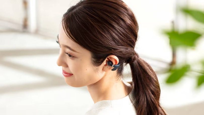 Vue de profil d'une jeune femme portant un écouteur Casio earU ER-100 en finition Navy Black au format open-ear cuff pincé autour du pavillon de l'oreille gauche, sans obstruction du conduit auditif, lumière naturelle douce et arrière-plan végétal flouté