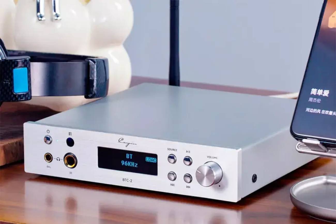 Façade du Cayin BTC-2 en finition argent montrant la sortie casque symétrique 4,4 mm (BAL) et la sortie asymétrique 6,35 mm (SE), l'écran OLED affichant BT 96KHz LDAC, les boutons SOURCE et lecture et le bouton de volume, avec un casque et un smartphone en lecture à droite
